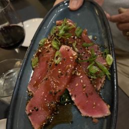 Togarashi Seared Ahi Tuna - A seared tuna appetizer with sliced tuna, garnished with chopped green onions, sesame seeds, and a drizzle of sauce, served on a black plate.