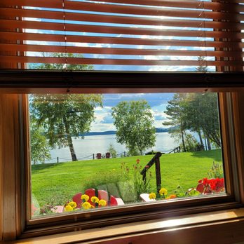 Loon Lodge - View from Lakeside One! - A view of a lake with trees, grass, and Adirondack chairs seen through a window with wooden blinds, capturing a sunny day. - Bed & Breakfast Near Me - Rangeley, Maine