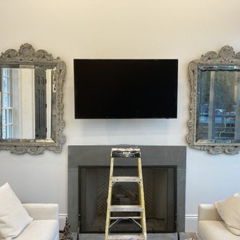 Fix It Eazy - Tv - A living room wall displays two large, ornate mirrors with carved frames on either side of a mounted flat-screen TV. Below the TV is a fireplace with a gray surround, and a small ladder is positioned in front of the fireplace. The room includes light-colored sofas with pillows, and a window reflection can be seen in the mirrors. - TV Mounting Near Me