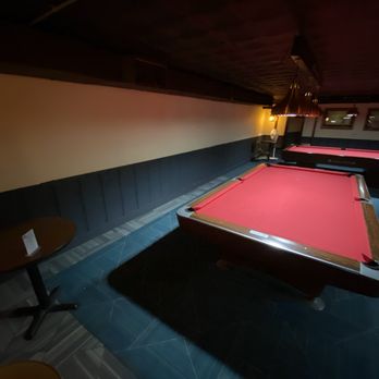 Chicago Billiard Cafe - The image shows a billiard hall with at least three pool tables with red felt surfaces, arranged in a row. The room has low lighting, creating a subdued atmosphere. A small round table with chairs is positioned against the wall on the left side. The ceiling is dark, and there are windows or framed pictures on the far wall. The overall setting suggests a relaxed environment for playing billiards. - Pool Halls Near Me - Chicago, Illinois