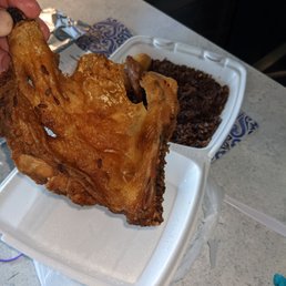 Fried chicken - A hand holds a piece of fried chicken over a takeout container filled with rice and black beans, on a kitchen counter.