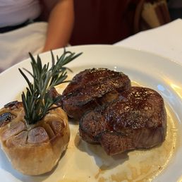 Filet Mignon Sliders - A white plate holds two cooked filet mignon steaks with a seared exterior, placed next to a roasted garlic head garnished with a sprig of rosemary. The dish is served in a restaurant environment, with a person partially visible in the background. The steaks appear to be seasoned and cooked to a medium or medium-rare level, with juices visible on the plate.