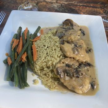 Kalamata - Chicken Marcella with rice, pilaf, and fresh vegetables. - A square white plate contains chicken covered in mushroom sauce, accompanied by a serving of rice and green beans with shredded carrots. The dish is set on a wooden table with a glass of water and wine nearby, indicating a restaurant setting. - Mediterranean Near Me - Arnold, California