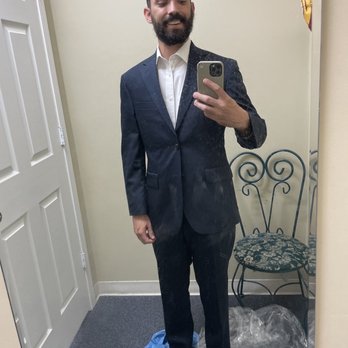 Grace Tailors - A man with a beard and glasses on his head is dressed in a dark suit and white shirt, taking a mirror selfie in a fitting room. The mirror is slightly dirty, and the background includes a chair with a floral cushion and some clothing or plastic on the floor. The setting appears to be a clothing store or fitting room. - Sewing & Alterations Near Me - Denver, Colorado