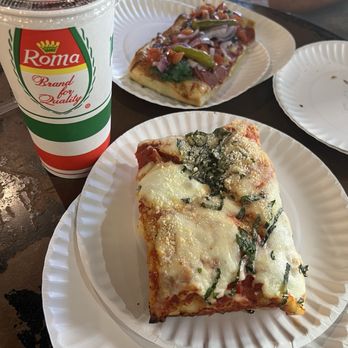 Slices - Sicilian tomato basil (bottom) & Chicago style hot dog (top) - A paper plate contains a slice of cheese and herb pizza, topped with cheese and chopped herbs. Another paper plate holds a rectangular pizza slice with toppings including green peppers, onions, and possibly bacon. A large soda in a Roma-branded cup with a straw is also on the table. The setting appears to be a casual dining environment with a dark table surface. - Pizza Near Me - Tempe, Arizona