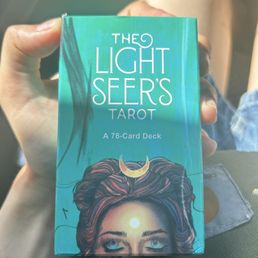 The image shows a person holding a box of 'The Light Seer's Tarot' deck, which features a vibrant, artistic illustration of a woman with flowing hair and a crescent moon on her forehead. The box indicates it contains 78 tarot cards and is designed for tarot reading and divination. The person's hand and part of their leg are visible, with natural lighting highlighting the colorful artwork on the box.