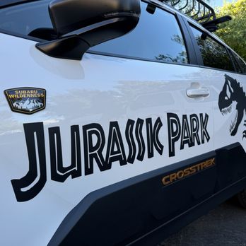 Big Boy Wraps - Jurassic Rides by Big Boy Wraps - A white Subaru Crosstrek displays Jurassic Park-themed decals, including a large 'JURASSIC PARK' logo and a dinosaur graphic on the side. The vehicle has a Subaru Wilderness badge near the front door and is parked outdoors with trees and sunlight visible in the background. The car's windows are tinted, and it has black wheels and side mirrors. - Vehicle Wraps Near Me - Martinez, California