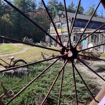 Three Sisters Sanctuary - Art - A rusty bicycle wheel is in the foreground, framing a rustic art building with sculptures and greenery in the background. - Parks Near Me - Goshen, Massachusetts