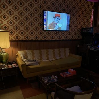 Thunderbird Lounge - The image shows a lounge with a yellow mid-century modern sofa covered with a zigzag patterned blanket. A wooden coffee table holds board games and magazines. To the left, a green lamp illuminates the space, and a small side table has phones and magazines. A flat-screen TV mounted on the wall displays a cartoon character wearing a hat and reading a paper. The wall behind the TV has retro geometric wallpaper, and the floor has a checkered tile pattern. The overall setting suggests a vintage or retro-themed lounge area. - Lounges Near Me - Phoenix, Arizona