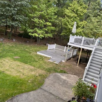 Concrete Solutions & Waterproofing - Before photo - The image shows a backyard with a wooden deck and stairs leading down to a grassy lawn. The deck has a white railing and an umbrella, with some outdoor furniture visible. The lawn is bordered by trees and bushes, providing a natural backdrop. A concrete pathway curves through the yard, and there are potted plants near the house wall. The scene appears to be a residential outdoor space with typical yard features. - Masonry/Concrete Near Me - Braintree, Massachusetts