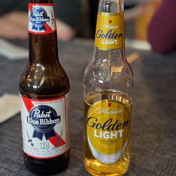 The Brass Lantern - beers - Two beer bottles, Pabst Blue Ribbon and Michelob Golden Light, are placed on a textured table surface in a casual setting. - Pubs Near Me - Chippewa Falls, Wisconsin