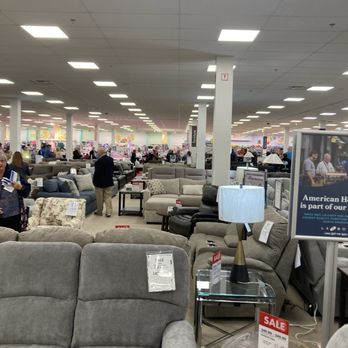 Boscov's - The photo depicts the interior of a large furniture store, featuring numerous sofas and tables arranged in rows. Sale signs are visible on some furniture pieces, and customers are browsing and walking through the store. The ceiling has evenly spaced fluorescent lights, and the store appears spacious and well-lit. - Department Stores Near Me - Rochester, New York