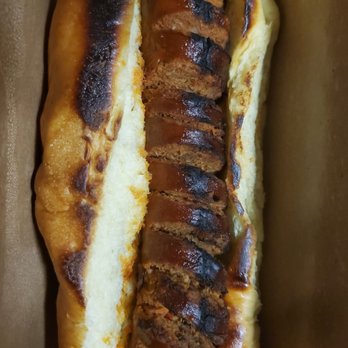 Fancy Franks - Chorizo sausage - A hot dog bun holds sliced, grilled chorizo sausage with visible grill marks and a slightly charred surface. The sausage slices are arranged in the bun, which appears soft and lightly toasted. The food is presented in a brown paper container, typical of casual food service settings. - Hot Dogs Near Me - Toronto, Ontario