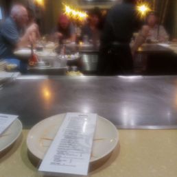 At the table - A blurry image shows diners seated at a teppanyaki grill with a chef standing in front of them, and a menu on the counter.