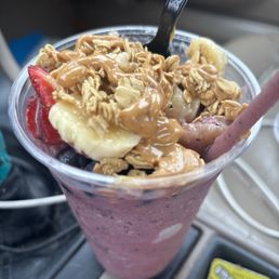 A berry smoothie served in a clear plastic cup, topped with granola, banana slices, and strawberries. A spoon and a pink straw are inserted into the drink. The background shows part of a vehicle interior and a yellow lighter on a surface.