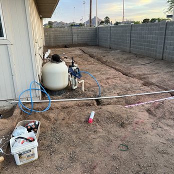 Divine Design Landscaping - A backyard area shows exposed dirt and construction pipes, with a large white water tank and various tools and supplies, including a plastic container with tools and a pink-handled tool on the ground. The area is enclosed by a gray concrete block wall, and the house's exterior wall is visible on the left. The scene suggests ongoing landscaping, irrigation, or hardscape installation work. - Landscaping Near Me - Phoenix, Arizona