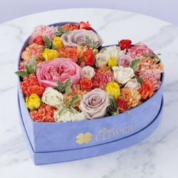 Pink Clover - Heart-shaped periwinkle box with a bright mix of roses, spray roses and carnations in pink, orange and yellow.