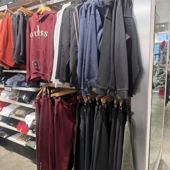 Guess - The image shows a retail store display with a variety of hoodies hanging on a metal rack, including red, black, gray, and blue options. Below, sweatpants in matching colors are hung on hooks. To the left, shelves contain folded clothing items, with some marked as on sale. The store has an industrial ceiling with exposed ductwork and lighting fixtures, and a glass wall or door is visible on the right side. - Men's Clothing Near Me - Paramus, New Jersey