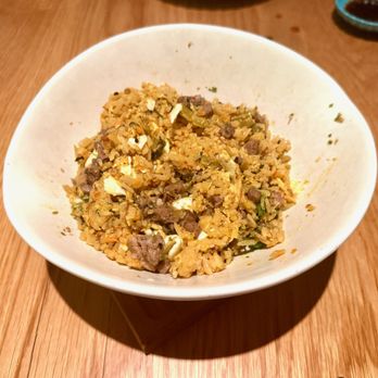 Makoto Vail - Wagyu Fried Rice (after mixing) - A ceramic bowl filled with fried rice featuring diced beef, chopped vegetables, and scrambled egg pieces, placed on a wooden table. The dish appears to be part of Japanese cuisine, possibly served at a restaurant specializing in sushi and Japanese dishes. In the background, a small dish of soy sauce and a blue bowl are partially visible. - Sushi Bars Near Me - Vail, Colorado