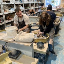 Private lesson. - Multiple people are working on pottery projects in a ceramic studio, using pottery wheels and clay. Shelves filled with pottery supplies, tools, and materials line the background. The studio has a bright window, and the workspace includes various pottery equipment and work surfaces.