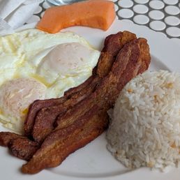 Garlic Rice - A breakfast plate includes two fried eggs with runny yolks, several strips of crispy bacon, a rounded serving of steamed white rice, and a slice of cantaloupe melon. The plate is set on a table with a patterned surface, and utensils are visible nearby.