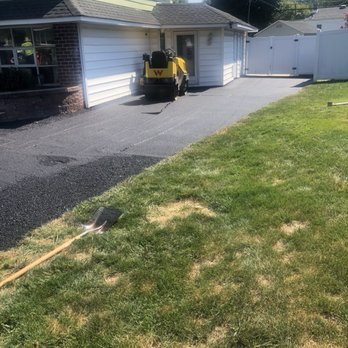 AS Paving - The image shows a small yellow construction vehicle parked on a newly paved black asphalt driveway adjacent to a house with white siding and a small porch. A shovel is lying on the grass nearby, and a white fence with a gate is visible in the background. The scene appears to be in a residential area during daytime. - Masonry/Concrete Near Me