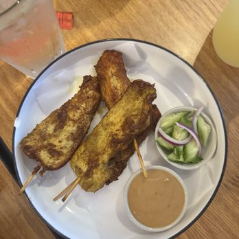 Paula's Thai Kitchen - Chicken satay - A plate with three skewered fried fish fillets, a small bowl of dipping sauce, and a cucumber salad with red onion slices. - Thai Near Me - Chicago, Illinois