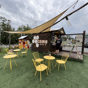 Comb Coffee - Seating area - The image shows an outdoor seating area with several yellow metal tables and chairs placed on artificial grass. A small wooden coffee stand labeled COMB Coffee Co. is situated in the background, with a person ordering at the window. There are decorative elements like a string of lights, a shade sail, and a small colorful shed nearby. The setting appears to be in a commercial area with a parking lot and other buildings visible in the background. - Coffee & Tea Near Me - Topton, Pennsylvania