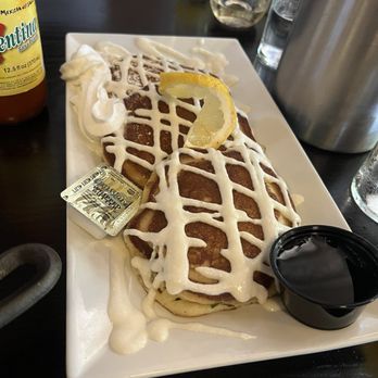 Port and Park - Lemon Ricotta Pancakes - A serving of two stacked pancakes topped with white icing drizzle, garnished with a lemon wedge, and accompanied by a small container of syrup. The pancakes are presented on a white rectangular plate, with a dollop of whipped cream and a packet of butter nearby. The background includes glasses, a metal container, and a bottle of hot sauce, indicating a breakfast or brunch setting. - New American Near Me - Chicago, Illinois
