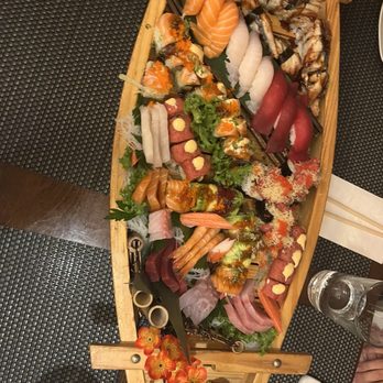 Nori - Super Boat - A large wooden sushi platter displays an assortment of sushi and sashimi, including slices of salmon, tuna, eel, and various rolls topped with fish roe and sauces. The platter is decorated with green leaves, shredded daikon, and edible flowers, and is placed on a dark textured table mat. There are glasses of water, chopsticks, and a smartphone nearby, indicating a dining setting. - Japanese Near Me - Montclair, New Jersey