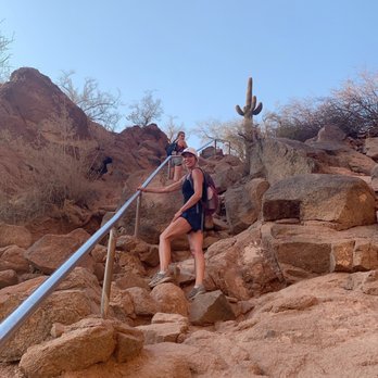 Echo Canyon Trail - A woman wearing a hat, sunglasses, and a backpack is ascending a rocky trail with a metal handrail in a desert landscape. The terrain is composed of reddish-brown rocks and sparse desert vegetation, including a cactus visible in the background. The sky is clear and blue, indicating a sunny day. Another hiker can be seen further up the trail. - Hiking Near Me - Phoenix, Arizona