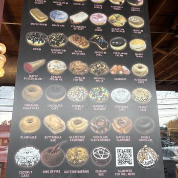 Voodoo Doughnut - A vertical menu board showcases an assortment of donuts with creative names and images, including flavors like 'Raised Glazed,' 'Chocolate Ring,' 'Chuckles,' and 'Voodoo Bubble.' The menu also lists vegan options and features donuts with themes such as 'Maple Bar,' 'Grape Ape,' 'Strawberry Go-Tart,' and 'Blueberry Cake.' The board includes a QR code for the full menu and a logo for the business, Voodoo Doughnut, located in Austin, TX. - Donuts Near Me - Austin, Texas
