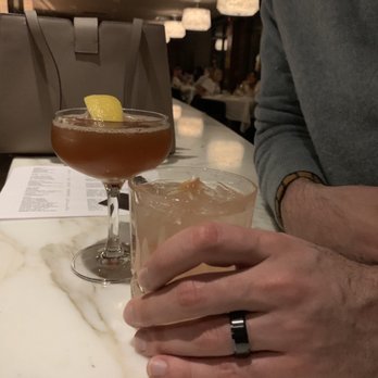 Forest - Two cocktails with ice are on a marble table in a restaurant, one garnished with a lemon wedge, with a person’s hand and arm visible. - New American Near Me - Birmingham, Michigan