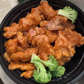 Lily’s Kitchen - Orange Chicken - A black takeout container holds orange chicken pieces coated in sauce, garnished with broccoli florets, on a light-colored surface. - Chinese Near Me - Malvern, Pennsylvania