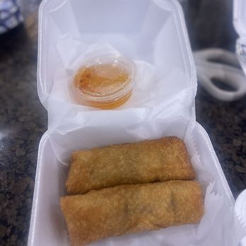 Thai Bistro - Pork Egg Roll - Two fried egg rolls are arranged side by side inside a white takeout container lined with paper. A small, clear plastic cup of dipping sauce is positioned above the egg rolls. The container is open, revealing the food, and the background includes a dark countertop with various jars and objects. - Thai Near Me - Brandon, Mississippi