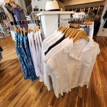 AZURI - A clothing rack in a boutique features white, black, and patterned garments on wooden hangers, including shirts and dresses. A white hat rests on a white display stand behind the rack. The store has wooden flooring, with additional clothing displays and customers visible in the background. The space is well-lit with ceiling spotlights, and the overall setting suggests a retail environment focused on women's fashion. - Clothing Rental Near Me - Delray Beach, Florida