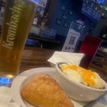 The Globe Pub - Shepherd's Pie - The image shows a bar counter with a tall glass of Kronenbourg beer, a bread roll on a small plate, and a bowl of mashed potatoes topped with melted cheese. In the background, there are bar decorations, a QR code stand, and a TV screen. The setting appears to be a casual pub or sports bar environment. - Sports Bars Near Me - Chicago, Illinois