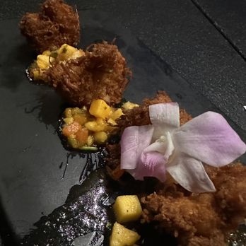 Fuse'd - Island Shrimp - The image shows a black plate with fried food items, possibly crab cakes or similar, garnished with a pink and white flower. There is a vegetable relish made of diced corn, carrots, and other vegetables, along with small yellow potato pieces. The presentation emphasizes a modern dining style with attention to visual detail and global culinary influences. - New American Near Me - Plainfield, Illinois