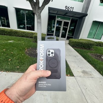 Rokform - A hand holding a Rokform rugged iPhone 15 Pro Max case box in front of a contemporary office building with large windows and a glass entrance. - Mobile Phone Accessories Near Me - Irvine, California
