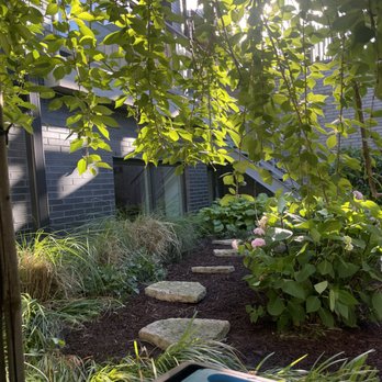 Chicago Landscaping - 10'X23' Planter & Deck, Finished Service - A garden with a stone pathway, lush plants, and flowering bushes, viewed from a shaded area with sunlight filtering through leaves. - Landscaping Near Me