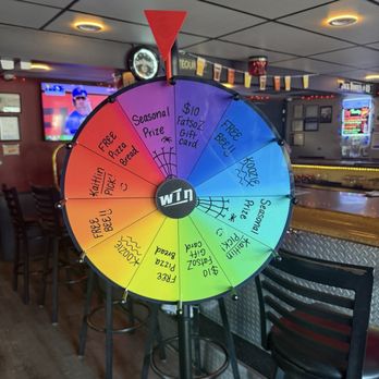Fatsoz Bar and Grill - spin the wheel football games - A multi-colored prize wheel is positioned inside a bar, featuring sections labeled with offers such as free pizza, a seasonal prize, a gift card, and free beer. The wheel is mounted on a stand, with a red triangular marker at the top. The background shows bar seating, a television screen, and bar decor, indicating a lively social environment. - Bars Near Me - Elgin, Illinois