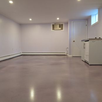 AD Technology - A large, empty room with light purple walls and a smooth, reflective floor. There is a small window near the ceiling and a portable air conditioning unit installed in the wall. A white door and a small white cabinet are also visible. The room appears to be a basement or storage area with minimal furnishings and lighting from ceiling fixtures. - Flooring Near Me