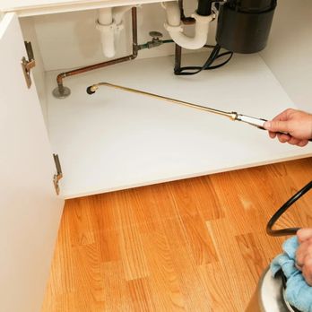 Safe Haven Pest Control - A pest control professional uses a long, thin wand to spray insecticide in the plumbing cabinet under a kitchen sink. - Pest Control Near Me Safe Haven Pest Control - A pest control professional uses a long, thin wand to spray insecticide in the plumbing cabinet under a kitchen sink. - Pest Control Near Me