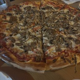 A large, round pizza topped with sliced mushrooms, crumbled sausage, melted cheese, and tomato sauce is displayed on a wooden table. The pizza is cut into slices, with some slices slightly separated. In the background, there is a glass of a light-colored beverage, a napkin, and utensils, indicating a dining setting. The pizza appears freshly baked with a golden crust.