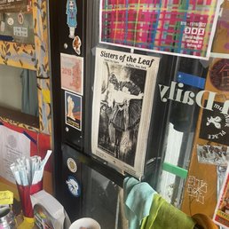 A black display case features stickers, posters, and a flyer for Sisters of the Leaf, with a colorful poster above and various items around it.
