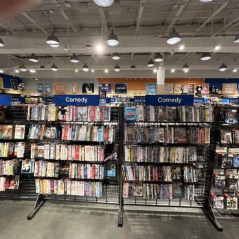 Fye - DVDs - The image shows multiple black metal racks filled with DVDs arranged in a retail store. The racks are labeled with genre categories such as Comedy, Horror, and Sci-Fi. The store has a high ceiling with numerous ceiling lights, and shelves with boxed items are visible in the background. The DVDs are tightly packed, and the store appears well-organized for browsing. - Music & DVDs Near Me - Gurnee, Illinois