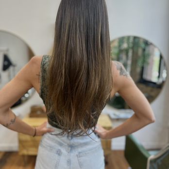 Salon Snob - A woman with long, straight brown hair is seen from behind, standing in a salon with her hands on her hips. She wears a sleeveless top and light-colored jeans. The salon features a mirror, a wooden table, and a green chair, with a window showing trees outside. The focus is on her hair and the salon environment. - Hair Stylists Near Me - Chicago, Illinois