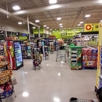 FreshCo - An indoor grocery store aisle features shelves stocked with snacks, drinks, and household items. The ceiling has exposed pipes and bright overhead lighting. A shopping cart is positioned in the middle of the aisle, and a child in a stroller is visible at the bottom right corner. The store has green signage and a clean, polished floor. - Grocery Near Me - Pickering, Ontario