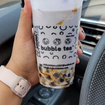Vivi Bubble Tea - Taro Milk Tea - A person is holding a bubble tea cup with tapioca pearls at the bottom inside a car. The cup has a printed design with cartoon faces and contact information. The vehicle's interior, including the dashboard and steering wheel, is visible, and the background shows part of a side mirror and outside scenery. - Bubble Tea Near Me - Columbus, Ohio