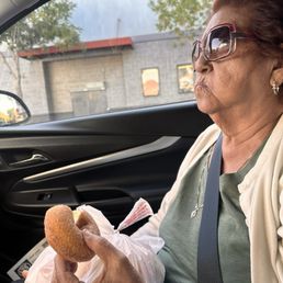 Churro donut. Mom approved! - A woman with short, reddish hair, wearing large sunglasses, earrings, and a light-colored cardigan, is seated in the passenger seat of a car. She is holding a donut in her right hand, which is partially wrapped in a paper bag. The car's interior is visible, with a window showing a building and trees outside. The woman appears to be eating the donut while seated in the vehicle.